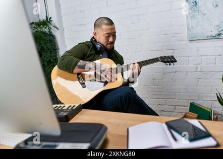 A young Asian man plays an acoustic guitar in his studio, surrounded by musical instruments and equipment. Stock Photo