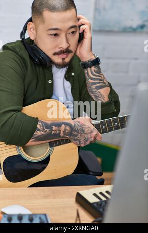 A tattooed Asian man in casual clothing plays an acoustic guitar while wearing headphones in his home recording studio. Stock Photo