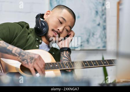 A young Asian man, wearing headphones, plays a guitar in his studio. Stock Photo