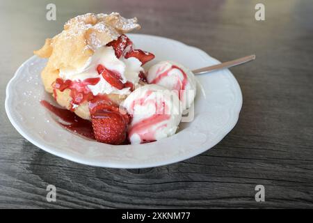 Cream puff filled with vanilla ice cream scoops, fresh strawberries and red fruit sauce, refreshing summer dessert on a plate and a gray wooden table Stock Photo
