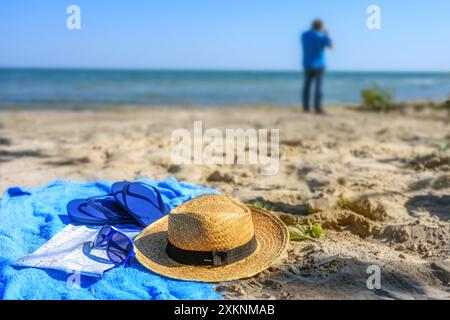 Straw hat, map, sunglasses and flip-flops on a blue towel on the beach and a blurred man in the background taking pictures of the sea, vacation photog Stock Photo