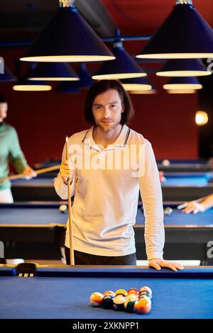 A man stands at a billiard table with his cue stick in hand, preparing to take a shot during a casual game with friends. Stock Photo