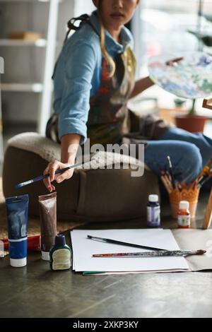 A young woman artist sits in her workshop, focused on her painting. Stock Photo