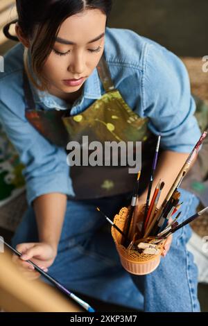 A young Asian woman artist is painting in her workshop, focusing intently on her work. Stock Photo