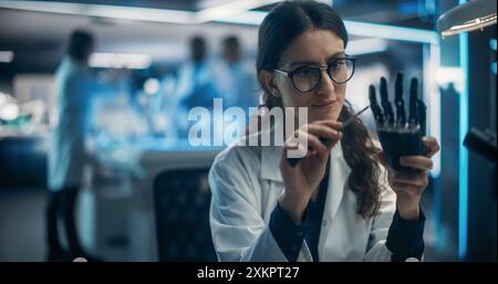 Beautiful Young Female Scientist Working on a Prosthetic Robotic Hand. On Background Diverse Team of Industrial Specialists Working on Advanced Remotely Controlled Mobility Robot Dog Stock Photo