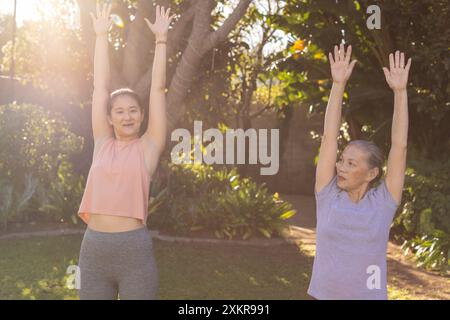 Exercising outdoors, asian grandmother and granddaughter stretching arms up in sunny garden Stock Photo