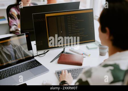 Over shoulder shot of male programmer surrounded by computers looking at screen with launched app while coding, his coworker sitting in front of him Stock Photo