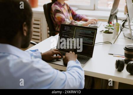 Over shoulder shot of African American man sitting at white wooden table and writing codes while his coworker typing on keyboard in blurred background Stock Photo