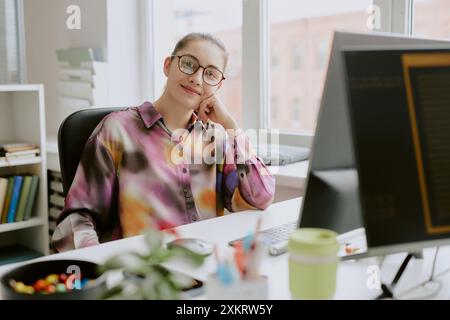 Portrait of young female programmer dressed in spotted blouse and wearing round glasses looking directly at camera and smiling sincerely while leaning Stock Photo