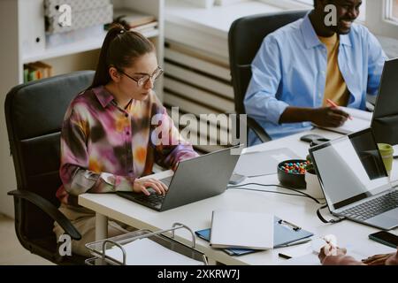 High angle shot of focused female worker sitting at table and typing keywords while her African American colleague writing report and smiling in blurr Stock Photo