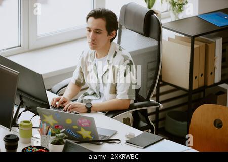 Young IT specialist typing on keyboard and looking at computer screen while working in cozy office surrounded by computers and thermal mugs of coffee Stock Photo