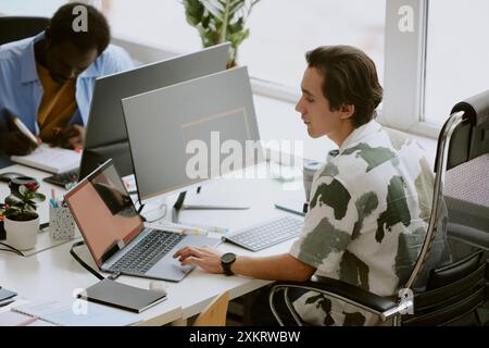 Multitask Caucasian programmer using gray laptop to search information while using computer to write code, his African American coworker writing repor Stock Photo
