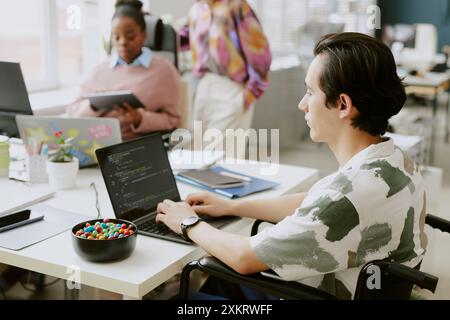 High angle shot of young IT specialist with disability writing code while his female coworkers working in blurred background Stock Photo