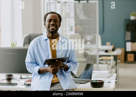 Young African American IT specialist holding silver touchpad and smiling widely while leaning on table in modern and comfortable office of high-end IT Stock Photo