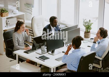 High angle shot of biracial team of operators with wired headphones and microphones on heads sitting at tables with computers on top in comfort office Stock Photo