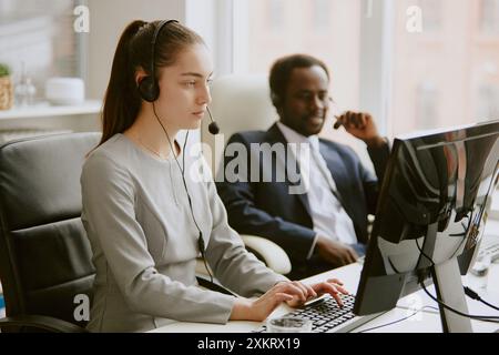Young Caucasian call center operator dressed in gray dress using computer while her African American male coworker helping client remotely Stock Photo