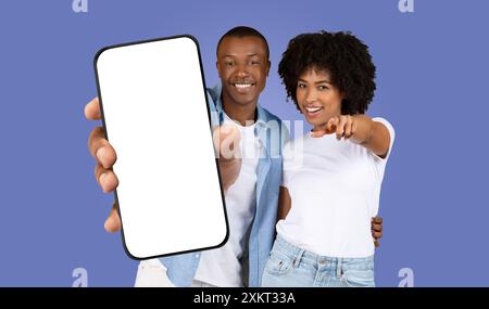 A young black couple stand together against a light blue background, smiling and pointing toward the camera. The man holds a smartphone with a blank w Stock Photo