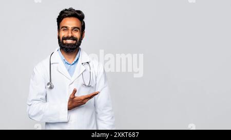A young Indian man with a beard, wearing a white lab coat and a stethoscope, stands in front of a gray background. He is smiling and gesturing with hi Stock Photo