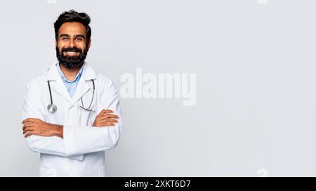 Indian man doctor with a dark beard and a white lab coat stands against a white background with his arms crossed and smiling at the camera. He is wear Stock Photo