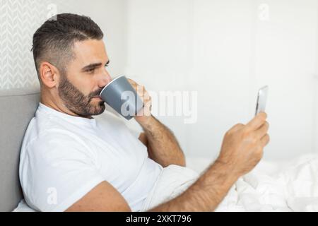 A man with a beard sits in bed, drinking from a gray mug and using his phone, with white bedding and a white wall behind him. Stock Photo