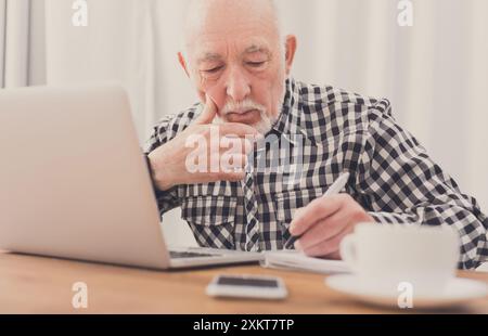 Mature man using laptop and writing in notepad at home desk. Education, recipe concept Stock Photo