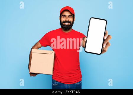 Indian delivery man wearing a red shirt and hat is holding brown cardboard box in his left hand and smartphone with blank screen in his right hand. He Stock Photo
