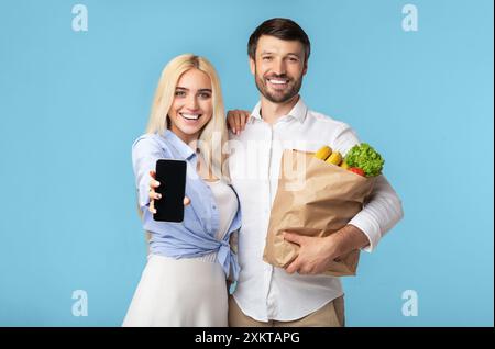 A young woman and man stand together, the woman holding a smartphone out toward the camera and the man holding a paper bag full of groceries. They are Stock Photo