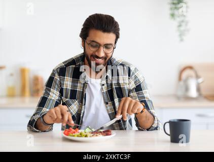 Positive arab guy hipster having healthy breakfast at home, eating fresh vegetables, sausages, drinking coffee, sitting at kitchen table, copy space. Stock Photo