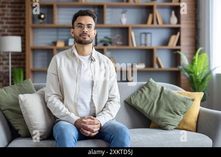 Confident young man sitting on sofa in modern living room with bookshelf in background. Casual attire and glasses, relaxed and thoughtful expression. Comfortable and stylish home setting. Stock Photo