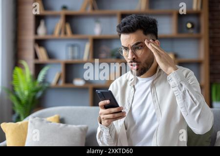 Young man in glasses looking surprised and shocked at his smartphone while sitting on couch at home. Expressing emotions, disbelief, and astonishment. Modern casual lifestyle concept. Stock Photo