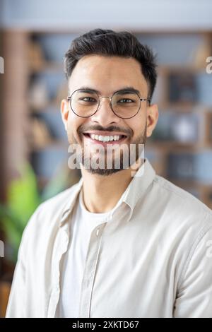 Smiling young man with glasses and beard wearing casual clothes indoors. Happy and confident expression, bright indoor setting, blurred background with bookshelves. Concept of positivity, youth, confidence, and casual style. Stock Photo