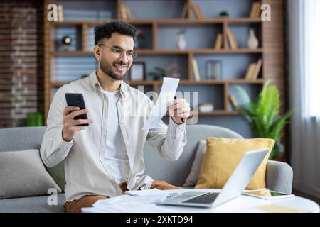 Smiling young man using smartphone and laptop to manage finances from home in cozy living room. Enthusiastic about going through bills and paperwork. Concept of home office, financial management Stock Photo