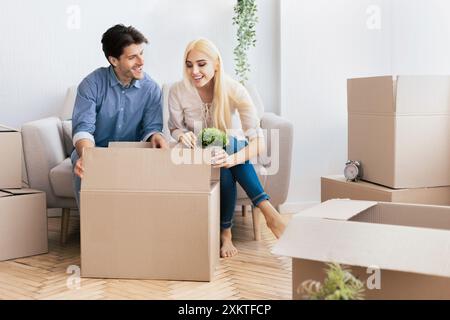 A young couple unpacks boxes in their new home, surrounded by other boxes and a plant. Stock Photo