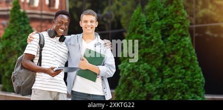 College Mates. Two multiethnic male students posing outdoors after classes, standing over university background, embracing and smiling at camera Stock Photo