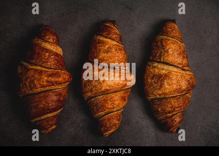 Three freshly baked croissants sit on a dark grey countertop, ready to be devoured. Their golden-brown crust glistens under the soft light, inviting a Stock Photo