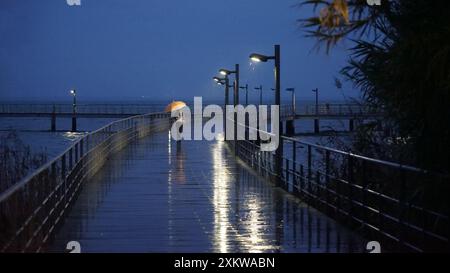 Pedestrian area with human figure with yellow umbrella on his back, on a rainy night with Vasco da Gama bridge located in Lisbon. Stock Photo