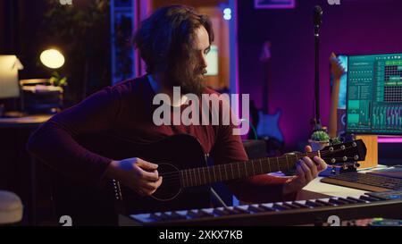 Portrait of male songwriter playing acoustic guitar and recording musical sounds to use for mix and master in home studio. Composing tunes with digital audio workstation software. Camera A. Stock Photo