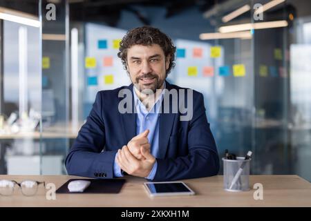 Confident businessman in modern office sitting at desk with digital tablet. Professional setting with glass walls and sticky notes on background. Ideal for business meeting Stock Photo