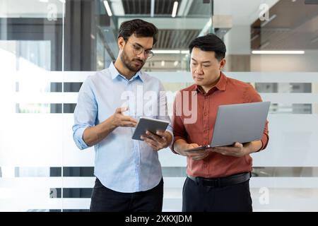 Two male office workers collaborate in modern office, using digital devices. Team discussion, sharing ideas, and analyzing data on laptop and tablet. Professional interaction business environment. Stock Photo