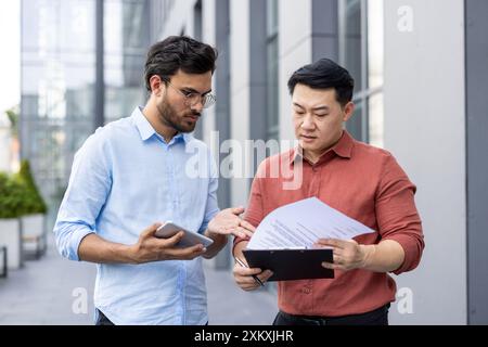 Two businessmen stand outside modern office building, engaged in conversation while reviewing documents and tablet. Concept of teamwork, collaboration, and communication in business environment. Stock Photo