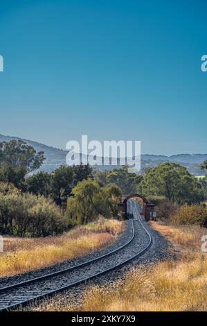 A grass-lined set of Railway lines lead viewer to an archway on a bridge over a tree-lined river in the small town of Woolbrook in New South Wales. Stock Photo