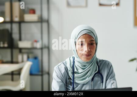 Young Muslim female virtual health assistant with stethoscope around neck looking at laptop screen during online medical consultation Stock Photo