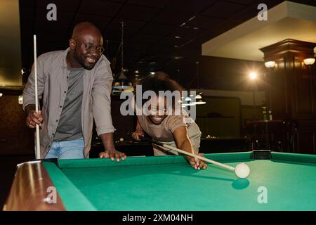 Happy young African American couple playing billiards wile smiling woman bending over green table and hitting white ball by cue Stock Photo