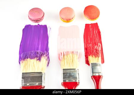 Three paintbrushes with red handles, each dipped in colorful paint, with matching macarons on a white background. Stock Photo