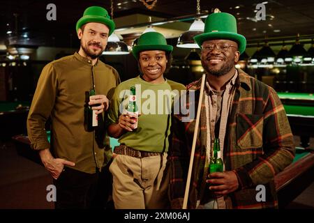 Three happy young friends in green hats and casual attire holding bottles of beer and looking at camera while standing in game room or pub Stock Photo