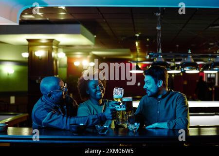 Three young cheerful friends in neon blue lit Irish pub sitting by bar counter and toasting with glasses of beer while enjoying party Stock Photo