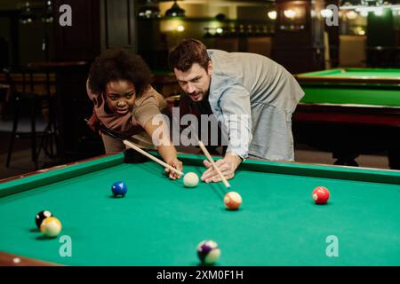 Young intercultural pool players with cues bending over wide billiards table with green top and looking at one of balls during game Stock Photo