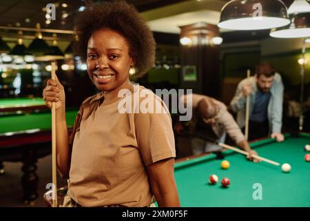 Young smiling female billiards player with cue in hand looking at camera against wide green table and two playing men in game room Stock Photo