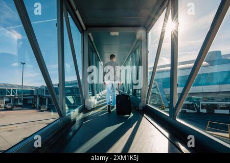 Travel by airplane. Rear view of man with hand baggage walking in passenger boarding bridge at airport on sunny day. Stock Photo