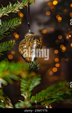 A copper golden Christmas bauble heart shaped ornament hangs on a pine branch, with blurred out Christmas lights in the background. Stock Photo
